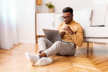 Afro Man Working On Laptop Having Coffee Sitting On Floor At Home. Freelance Career And Lifestyle. Selective Focus