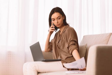 Belgeler İnceleniyor. Elinde kağıt tutan ve cep telefonuyla konuşan genç bir kadın. Ev ofisinde çalışıyor.