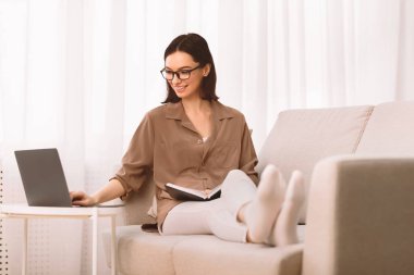 Bilgi kavramı. Gözlüklü rahat kız koltukta oturuyor, kitap okuyor, dizüstü bilgisayarı tutuyor ve kopyalama alanı kullanıyor.