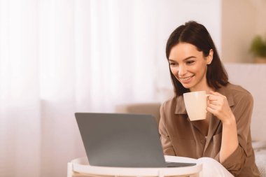 İnternette sörf yapmak. Mutlu kız bir fincan sıcak kahveyle rahatlıyor, dizüstü bilgisayar kullanıyor, evde vakit geçiriyor, boş zaman geçiriyor.