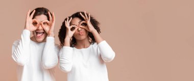 Funny multiracial friends fooling, making glasses with fingers, having fun together over yellow background