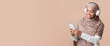 Cool music app. Smiling black muslim girl in hijab listening her favorite songs with wireless headphones online on smartphone
