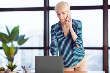 Business Calls. Entrepreneur Lady Talking On Mobile Phone Standing Near Laptop Computer In Modern Office. Selective Focus, Free Space