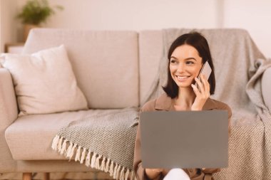Mutlu genç bir kadın cep telefonuyla konuşuyor evde yerde oturuyor, dizüstü bilgisayar kullanıyor, kopyalama alanı kullanıyor.