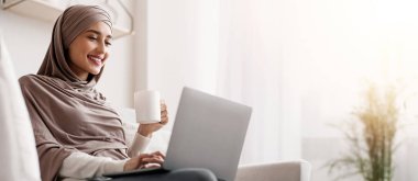 Ev Tatili. Genç Arap kadın tesettürde dizüstü bilgisayar kullanıyor ve kahve içiyor, internetten film izliyor, oturma odasında dinleniyor, boş yer arıyor.