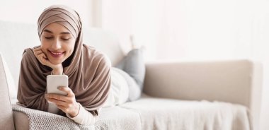 tesettürlü çekici Müslüman kadın evde rahat bir kanepede uzanırken akıllı telefon kullanıyor, sosyal ağlara mesaj atıyor ya da göz gezdiriyor, kapatıyor