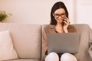 Kurumsal İletişim. Cep telefonuyla konuşan gülümseyen kız evde dizüstü bilgisayarda çalışıyor, koltukta oturuyor, kopyalama yapıyor.