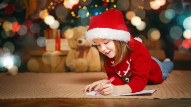 Kırmızı şapkalı şirin kız yere yatmış ve Noel Baba 'ya mektup yazıyor.