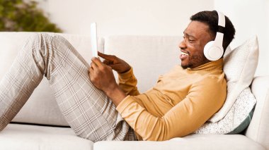 Hafta sonu konsepti. Kulaklık takan, Tablet Bilgisayar kullanan, Müzik dinleyen, koltukta uzanan mutlu Afrikalı Amerikalı adam.