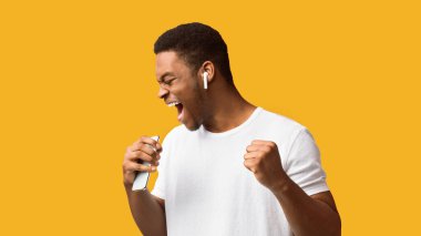 Neşe kavramı. Heyecanlı siyah adam en sevdiği şarkıyı söylüyor, akıllı telefonu mikrofon olarak kullanıyor, turuncu stüdyo arka planı.