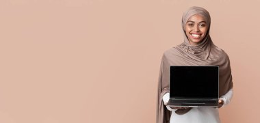 Cool Website. Smiling Black Muslim Girl In Hijab Holding Laptop Computer With Blank Black Screen For Mockup, Standing Over Yellow Background