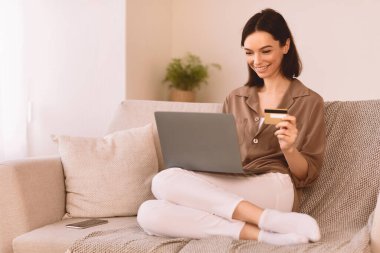 Çevrimiçi Mağaza. Mutlu kız internetten alışveriş yapıyor. Laptop ve kredi kartı kullanıyor. Kanepede oturuyor.