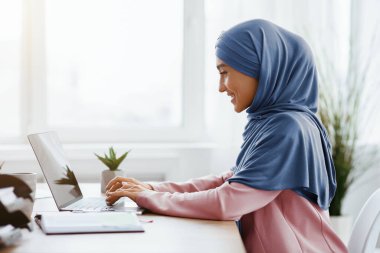 Ofis İslami Kadınlar için çalışıyor. Türbanlı Mutlu Müslüman Kız masadaki dizüstü bilgisayarda çalışıyor, klavyede yazıyor, boş alanla yan görünüm
