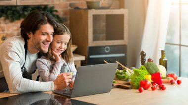 Mutfak Bloğu. Neşeli genç baba ve küçük kız mutfakta yemek pişirirken dizüstü bilgisayar kullanıyor, ev yapımı yemek tariflerini kontrol ediyor, internette geziniyor, evde eğleniyor, boş alan arıyor.