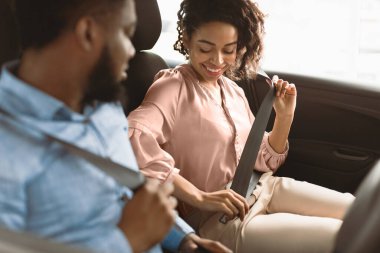 New Car Concept. Smiling Black Couple Putting On Seat Belts Testing Automobile In Luxury Dealership Showroom. Selective Focus