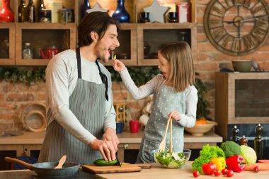 Genç, mutlu bir aile babası ve kızı mutfakta birlikte yemek pişirirken, küçük bir kız babasını sebzelerle beslerken, evde sağlıklı yiyecekler hazırlarken, öğle yemeği hazırlarken ya da akşam yemeği hazırlarken, ya da boş bir alanda,