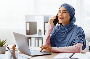 Cep telefonuyla konuşan ve modern ofiste dizüstü bilgisayar kullanan mutlu Müslüman iş kadını, İş yerinde Çağrı Olan Kadın Çalışan, Boş Alan