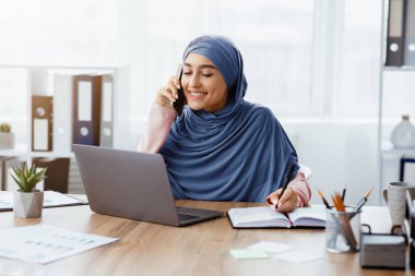 İş hayatı tarzı. Ofisteki meşgul Orta Doğulu iş kadınının portresi, cep telefonuyla konuşmak, dizüstü bilgisayar kullanmak ve not almak.