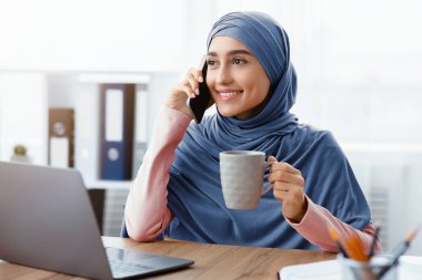 Gülümseyen Müslüman kadın yönetici cep telefonuyla konuşuyor ve işyerinde kahve içiyor.