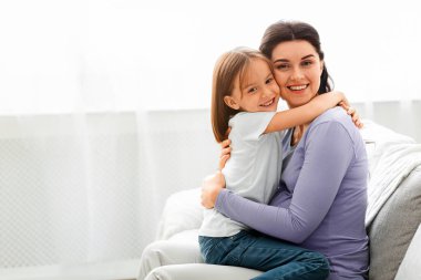 Sevgi dolu küçük bir kız. Genç annesini kucaklıyor. Kameraya gülümsüyor.