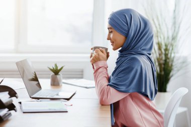 İşte mola. Genç Müslüman kadın girişimci işyerinde bilgisayarını, yan görüntüsünü ve boş alanı kullanarak kahve içiyor.