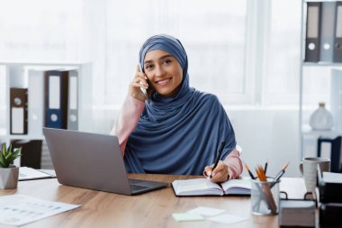 Modern Sekreter. Başörtülü Müslüman Kadın cep telefonuyla konuşuyor ve ofiste dizüstü bilgisayarla çalışırken not alıyor.