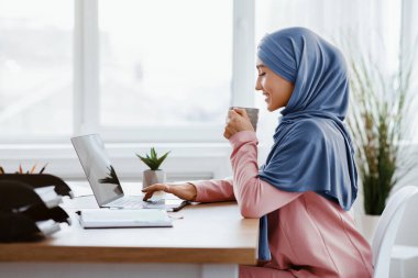 Genç Arap iş kadını, modern ofiste dizüstü bilgisayarla çalışırken masasında bir fincan kahvenin keyfini çıkarıyor.