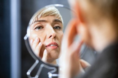 Cilt Yaşlanma Konsepti. Orta yaşlı kadın, yuvarlak aynada kırışıklıklara bakıyor. Yüzüne dokunuyor. Kapalı bir odada duruyor. Seçici Odaklanma