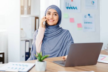 İş İletişimi. Modern ofiste çalışırken cep telefonuyla konuşan ve dizüstü bilgisayar kullanan neşeli bir Arap kadın.