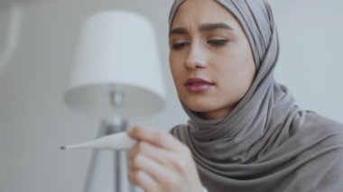 Bir kadın endişe ile bir termometre gözlemler, gri bir tesettür giyinmiş, onun yaşam alanında bir an kaygı yansıtır.