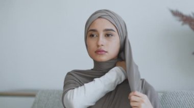 Bir kadın, yumuşak doğal ışıkla çağdaş bir odada otururken tesettürünü dikkatlice ayarlar..