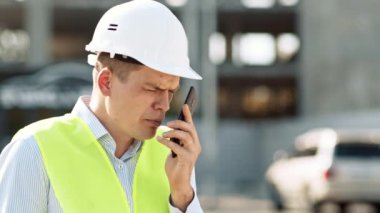 Bir inşaat işçisi, yoğun bir inşaat alanında çelik yelek ve kask giyerken cep telefonuyla iletişim kuruyor..
