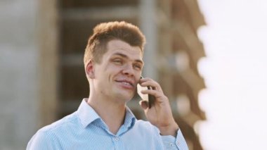 Gülümseyen genç bir adam, gün batımında bir inşaat alanının arka planına karşı bir telefon görüşmesi yapıyor..