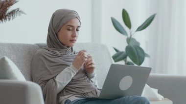 Gri tesettürlü bir kadın, iyi aydınlatılmış bir oturma odasında, dizüstü bilgisayarına dikkatlice bakıyor..
