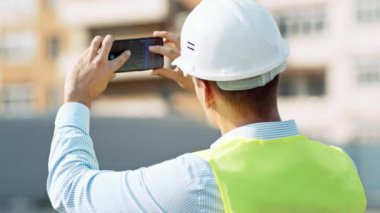 Kask ve güvenlik yeleği giyen bir inşaat işçisi akıllı telefonuyla bir anı yakalıyor..