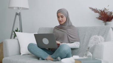 Dizüstü bilgisayarında tesettürlü bir kadın, parlak, şık bir oturma odasında rahat bir koltukta bacak bacak üstüne atmış..