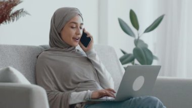 Tesettürlü genç bir kadın, yeşil bir oturma odasında dizüstü bilgisayarında yazarken bir telefon görüşmesi yapıyor..
