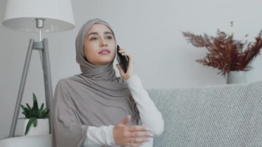 Gri tesettürlü bir kadın, çağdaş bir yaşam alanındaki bir kanepede dinlenirken telefon görüşmesi yapar..