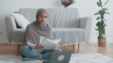 Türban takmış bir kadın, yumuşak mobilyalar ve parlak bir yaşam alanında dizüstü bilgisayarla yerde otururken kağıtları inceliyor..