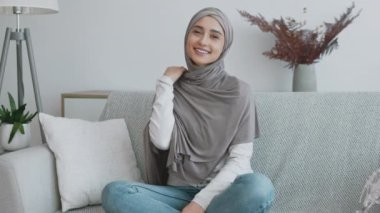 Başörtülü bir kadın rahat bir şekilde koltukta oturur, şık bir oturma odasında günlük kıyafetlerini giyer ve sıcacık gülümser..