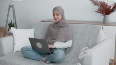 Müslüman bir kadın kanepede bağdaş kurarak oturur. Dizüstü bilgisayarına odaklanır. Bitkiler ve dekorlarla dolu rahat bir yaşam alanında..