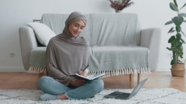 Müslüman bir kadın oturma odasında yerde oturmuş yanında dizüstü bilgisayarla gevşetici bir atmosferle not defteri yazıyor..
