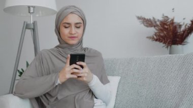 Gri tesettürlü bir kadın şık bir oturma odasında rahatça otururken akıllı telefonuyla yazı yazıyor..