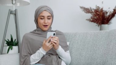 Başörtüsü takan bir kadın oturma odasında rahat bir kanepede otururken akıllı telefonuna bakarken şaşkınlığını dile getirir..