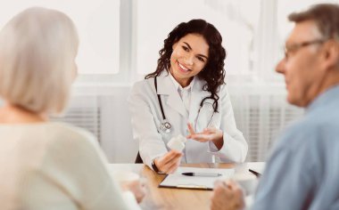 Visiting Doctor. Smiling latina physician showing mature couple pills, pointing at plastic bottle, giving prescription