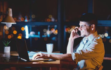 Erkek girişimci cep telefonuyla konuşuyor. Modern ofiste bütün gece dizüstü bilgisayarda çalışıyor. İş hayatı tarzı. Boşluğu Kopyala