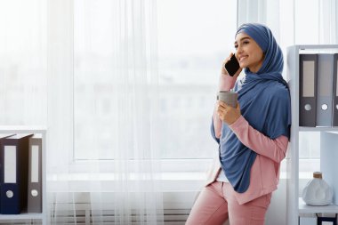 Genç Müslüman iş kadını ofiste akıllı telefon ve kahveyle dinleniyor, pencerenin yanında duruyor ve elinde sıcak içecekle fincanı tutuyor, fotokopi çekiyor.