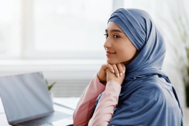 İş yerinde tesettür. Başörtüsü takan gülümseyen Müslüman iş kadınının profil portresi, işyerinde oturup başka yere bakmak, boşluğu kopyalamak.