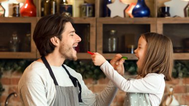Sevimli küçük kız ve genç babası mutfakta taze salata pişirirken birbirlerini sebzelerle besliyorlar.