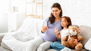 Evdeki kanepede otururken küçük kızıyla selfie çeken hamile bir kadın.